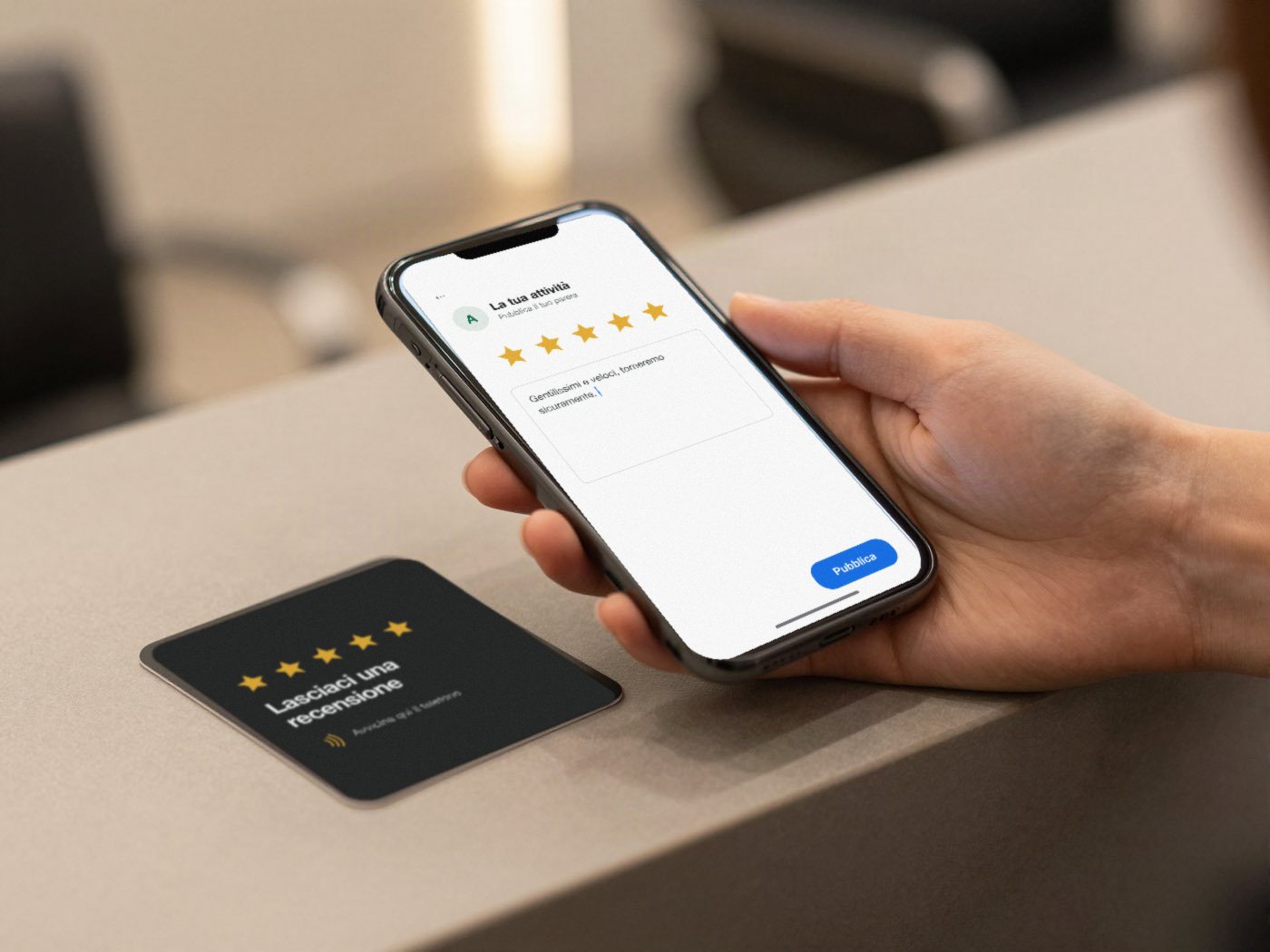 Una card scura con cinque stelle e la scritta Lasciaci una recensione, appoggiata sul bancone; accanto, una mano tiene il telefono con la finestra della recensione già aperta.